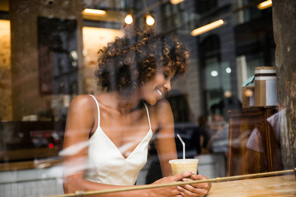 Beautiful afro woman sitting in a coffehouse - Pretty middle aged girl drinking coffee in a bar, reflection effect from window glass - 写真・画像