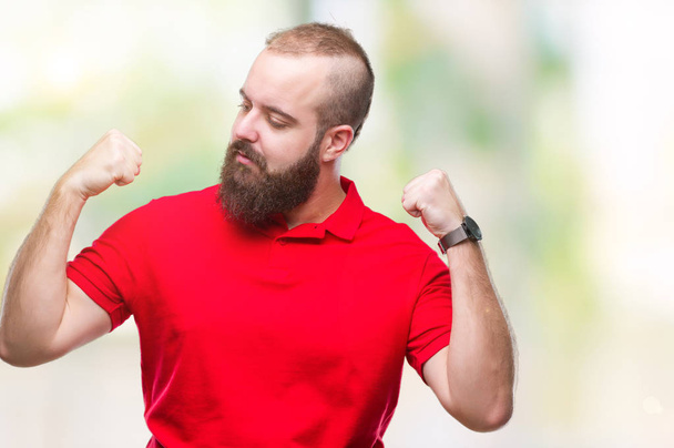 Молодой кавказский хипстер в красной рубашке на изолированном фоне показывает мышцы рук, гордо улыбающиеся. Фитнес-концепция
. - Фото, изображение