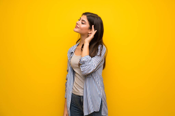Menina adolescente sobre a parede amarela ouvindo algo, colocando a mão na orelha
 - Foto, Imagem