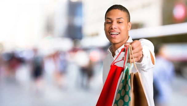 Νεαρός αφρικανική αμερικανική άνθρωπος με λευκό πουκάμισο έκπληκτος κρατώντας πολλές σακούλες ψώνια στο κέντρο της πόλης - Φωτογραφία, εικόνα