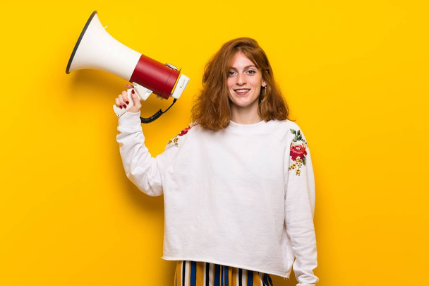 Young redhead woman over yellow wall holding a megaphone - Fotó, kép