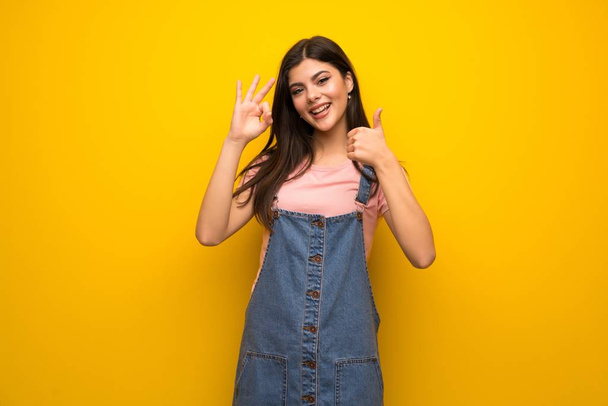 Tiener meisje over gele muur weergegeven: ok bord met en het geven van een duim omhoog gebaar - Foto, afbeelding