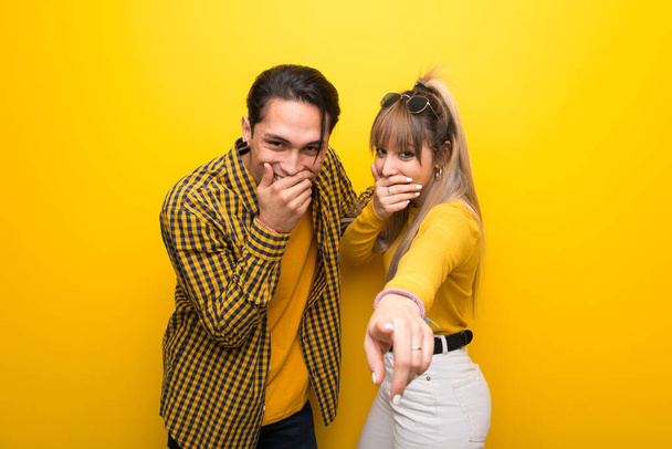 Νεαρό ζευγάρι πάνω από έντονο κίτρινο φόντο δείχνοντας με το δάχτυλο σε κάποιον και γελώντας - Φωτογραφία, εικόνα