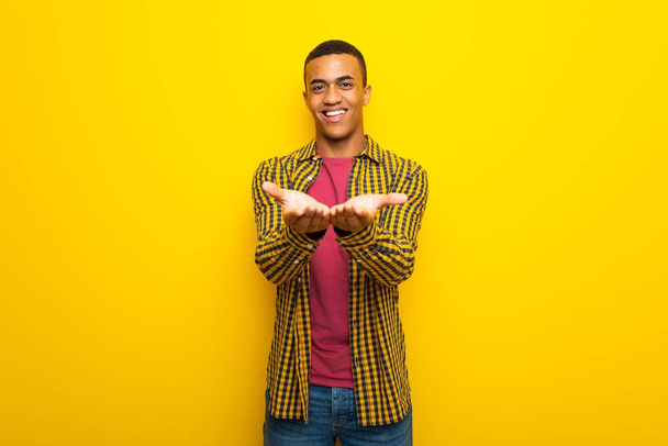 Afro Αμερικανός νεαρός σε κίτρινο φόντο κρατώντας copyspace φανταστικό στην παλάμη για να εισαγάγετε μια διαφήμιση - Φωτογραφία, εικόνα