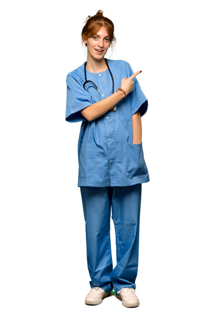 Ένα ολόσωμο πλάνο του μια νεαρή κοκκινομάλλα νοσοκόμα τοποθετώντας το δείκτη στην πλευρά να παρουσιάσει ένα προϊόν πέρα από το απομονωμένο λευκό φόντο - Φωτογραφία, εικόνα