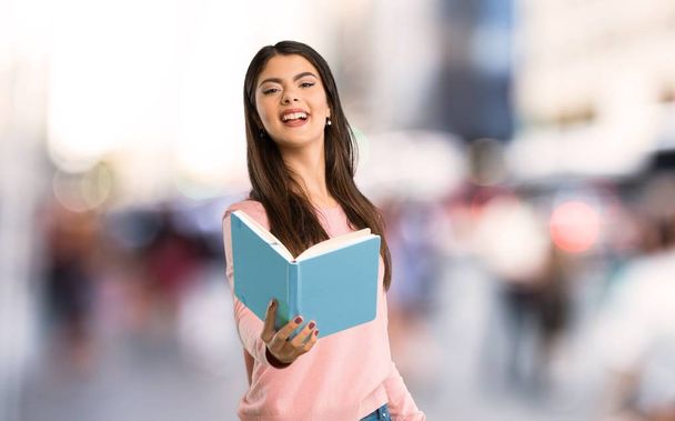Девочка-подросток в розовой рубашке держит книгу и дает ее кому-то в городе
 - Фото, изображение