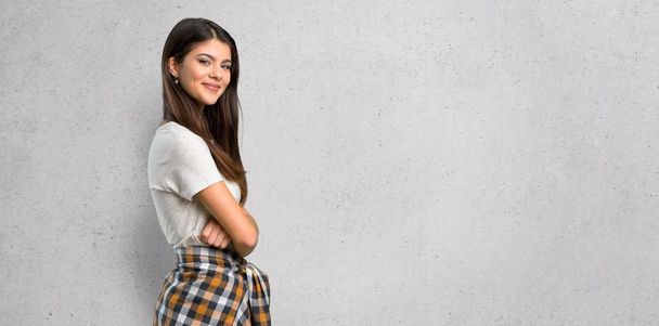 Teenager girl with shirt tied to the waist keeping the arms crossed in lateral position while smiling on textured wall background - Foto, Imagem