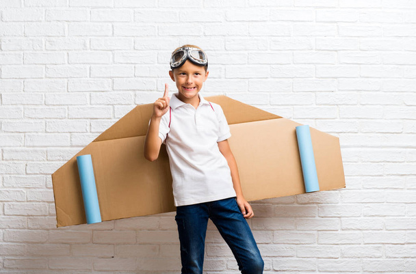 Junge spielt mit Flugzeugflügeln aus Pappe auf dem Rücken und zählt Zeichen Nummer eins - Foto, Bild