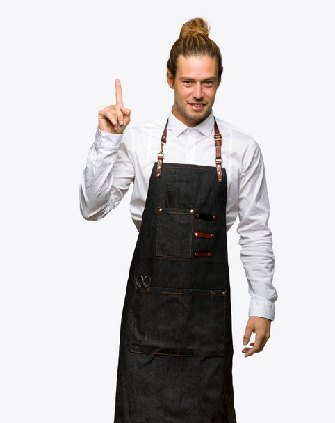 Barber man in an apron showing and lifting a finger in sign of the best on isolated background - Photo, Image