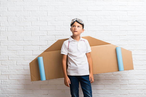 Jongen spelen met kartonnen vliegtuig vleugels op zijn rug twijfels en met confuse gezicht expressie - Foto, afbeelding