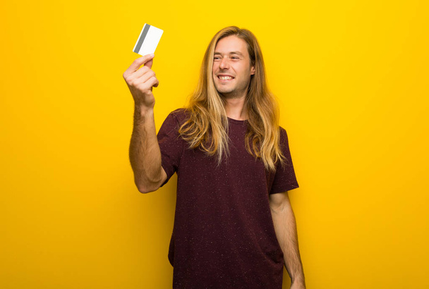 Blond man with long hair over yellow wall holding a credit card and thinking - Valokuva, kuva