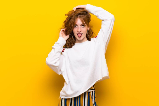 Jonge roodharige vrouw over gele muur tong tonen op de camera met grappige blik - Foto, afbeelding