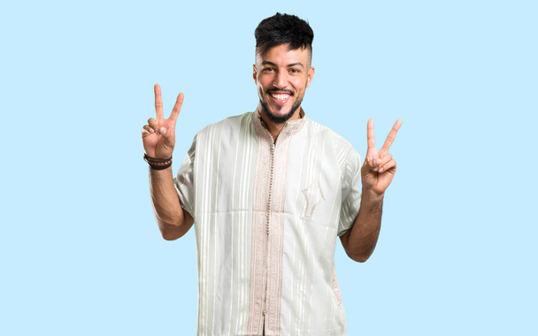 Arabic young man wearing typical arab clothes smiling and showing victory sign on blue background - Fotoğraf, Görsel