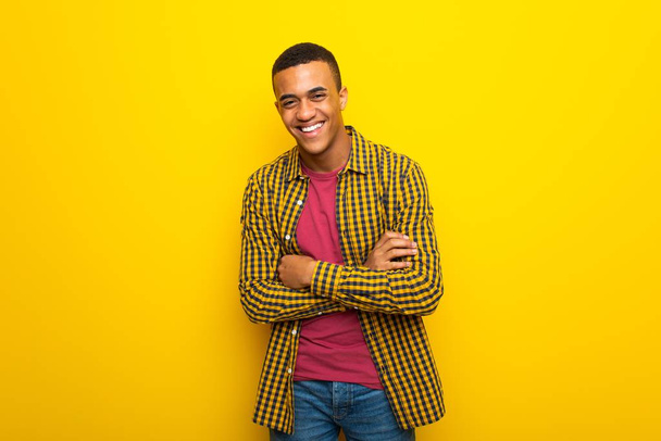 Young afro american man on yellow background keeping the arms crossed while smiling - 写真・画像