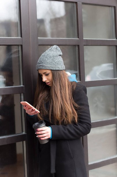νεαρή όμορφη γυναίκα καστανά μαλλιά γκρι πλεκτό καπέλο μαύρο παλτό με καφέ να πάει Κύπελλο και κινητό τηλέφωνο στο δρόμο - Φωτογραφία, εικόνα
