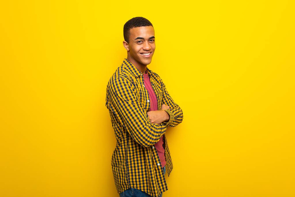 Young afro american man on yellow background keeping the arms crossed in lateral position while smiling - Fotó, kép