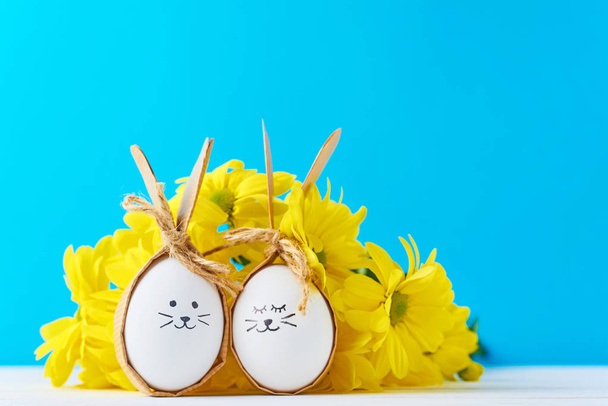 Two eggs with drawing faces with yellow flowers on a blue background - Fotoğraf, Görsel