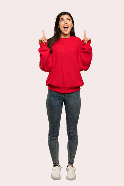 Een full-length shot van een tiener meisje met het rode trui wijzen omhoog en verbaasd over geïsoleerde achtergrond - Foto, afbeelding