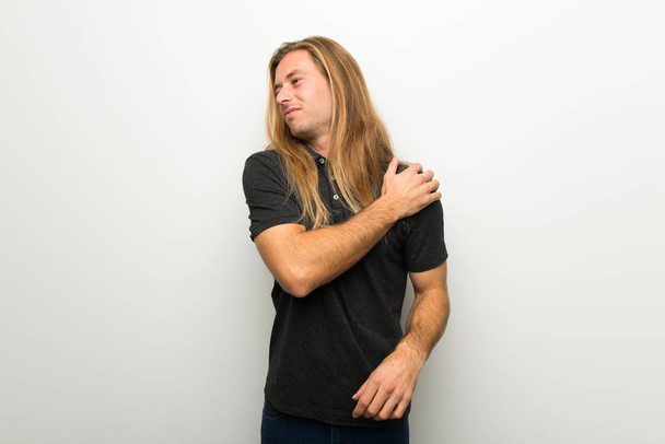 Ξανθός άντρας με μακριά μαλλιά σε λευκό τοίχο που πάσχουν από τον πόνο στον ώμο για να κάνει μια προσπάθεια - Φωτογραφία, εικόνα