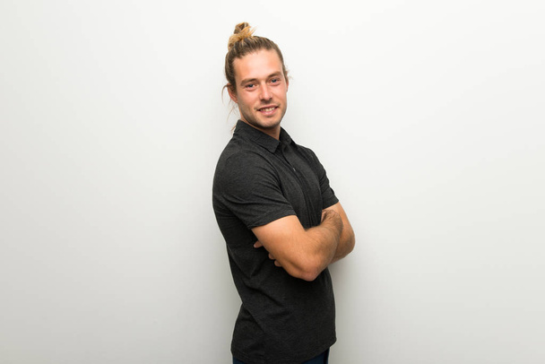 Blond man with long hair over white wall keeping the arms crossed in lateral position while smiling - Fotografie, Obrázek