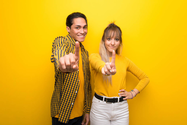 鮮やかな黄色の背景の上に若いカップルは、指を示し、持ち上げる - 写真・画像