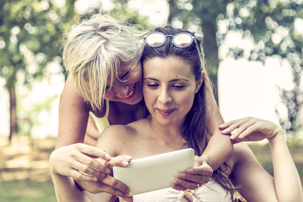 mother and daughter in swimsuit having fun watching a digital tablet in a nature park - Фото, изображение