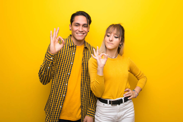 Mladý pár přes zářivý žlutý podklad ukazující na dobré znamení s prsty - Fotografie, Obrázek