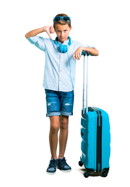 Full body of Kid with sunglasses and headphones traveling with his suitcase showing thumb down sign with negative expression on isolated white background - Photo, image