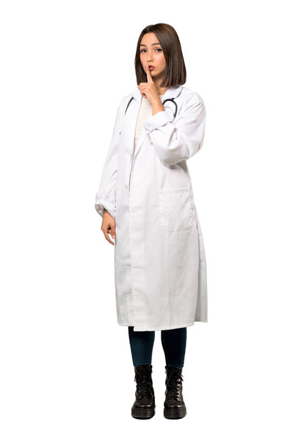 A full-length shot of a Young doctor woman showing a sign of silence gesture putting finger in mouth over isolated white background - Fotoğraf, Görsel