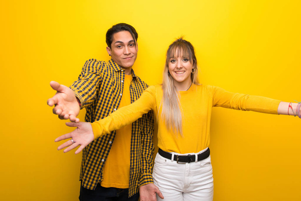 junges Paar vor lebendigem gelben Hintergrund präsentiert und lädt ein, mit der Hand zu kommen - Foto, Bild