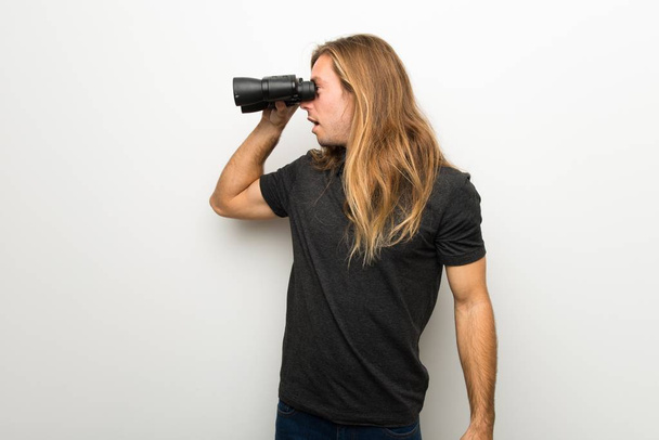 Blond man with long hair over white wall and looking in the distance with binoculars - Fotoğraf, Görsel