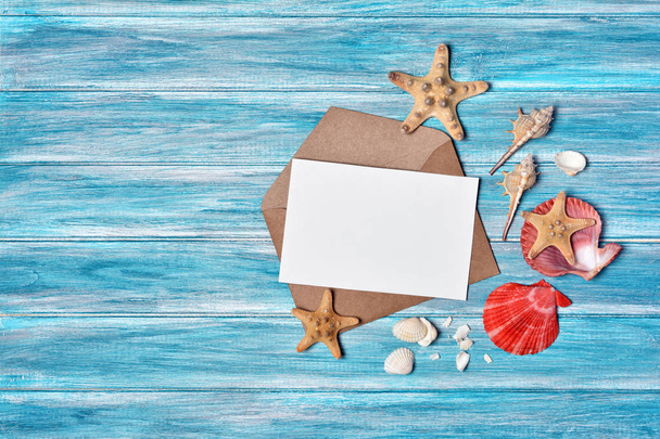Top view on mock-up of white paper card for photo or lettering on kraft envelope with starfish and seashells on blue wooden background with copy space - Foto, Imagen