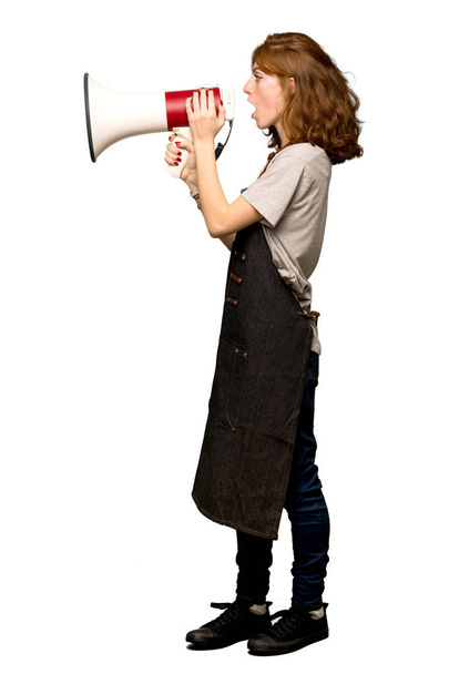 A full-length shot of a Young redhead woman with apron shouting through a megaphone over isolated white background - Fotografie, Obrázek