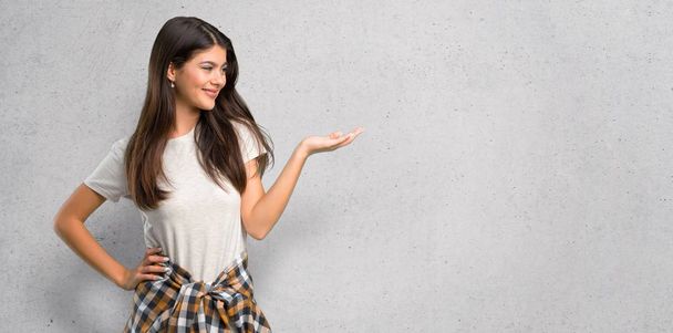 Teenager girl with shirt tied to the waist holding copyspace imaginary on the palm to insert an ad on textured wall background - Photo, Image
