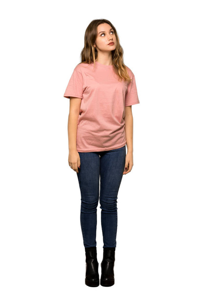 eine Ganzkörperaufnahme eines Teenagermädchens mit rosa Pullover, das mit der Hand auf das Kinn auf isoliertem weißem Hintergrund herabblickt - Foto, Bild