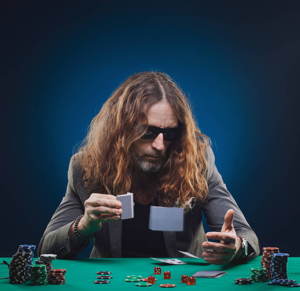 Long-haired handsome man playing poker in a casino. Studio concept on blue background - 写真・画像