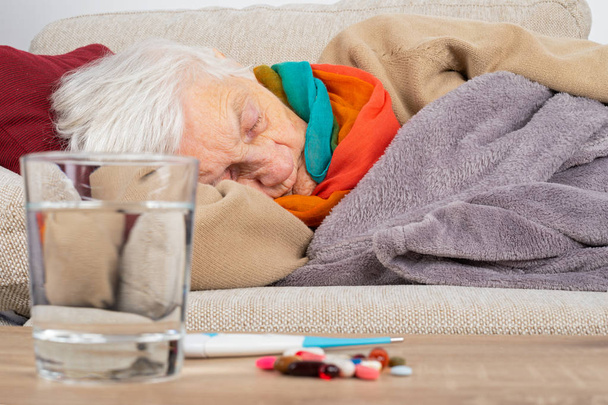 Senior woman feeling sick is lying on the couch wrapped in a blanket - Influenza, sore throat, headache - Medication, tissues and water in front of her - 写真・画像