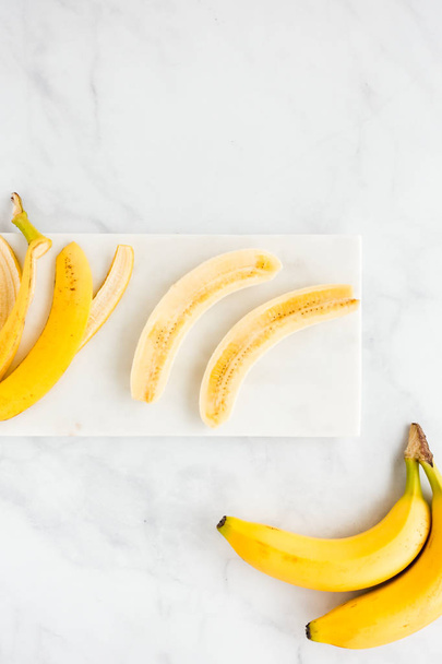 Banana peel and a peeled banana cut lengthwise in half on white marble board and background. Top view. - Photo, Image