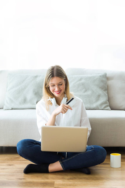 Tiro de mujer joven feliz sosteniendo la tarjeta de crédito blanca para ir de compras en línea con el ordenador mientras está sentado en el suelo en casa
. - Foto, Imagen