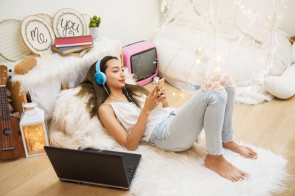 Atraktivní krásné asijské žena hrát sociální média, online nakupování, poslouchat hudební video smartphone a sluchátka v obýváku ozdobný. - Fotografie, Obrázek