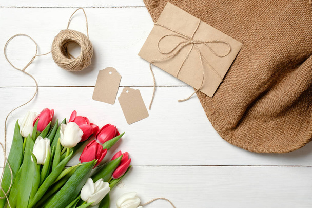 Bouquet of tulips flowers, kraft envelope, twine, tags, burlap on white wooden table. Vintage greeting card for womans day, mothers day, birthday, easter. Rustic background, flat lay, above view. - Foto, imagen