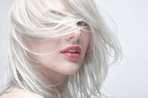 Portrait of a young beautiful blonde woman with plump red lips on a white background close up. Fashionable fancy hairstyle - 写真・画像