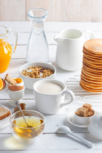 Healthy breakfast eating concept, various morning food - pancakes, soft-boiled egg, toast, oatmeal, granola, fruit, coffee, tea, orange juice, milk on white wooden table - Foto, Bild
