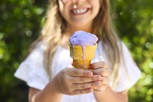 Portrait of the cute little girl with funny expression holding ice cream cone outside against bright nature background. Close up - Photo, Image