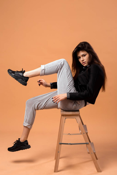 Dark-haired girl dressed in white shirt, gray trousers, black jacket  and black sneakers is sitting on a wooden stool on the beige background in the studio - Foto, Imagem
