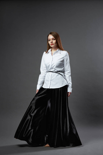 Beautiful young woman dressed in white shirt with black belt and wide black trousers stands on the dark gray background in the studio - Foto, Bild