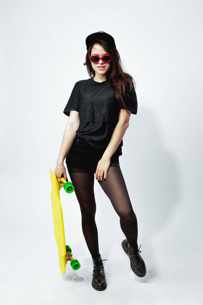 Funny young dark-haired girl in a black top, shorts, tights, cap and red sunglasses with yellow  skateboard is posing on the white background in the studio - Foto, Bild