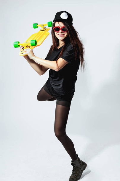 Happy young dark-haired girl in a black top, shorts, tights, cap and red sunglasses with yellow  skateboard is posing on the white background in the studio - Foto, Bild