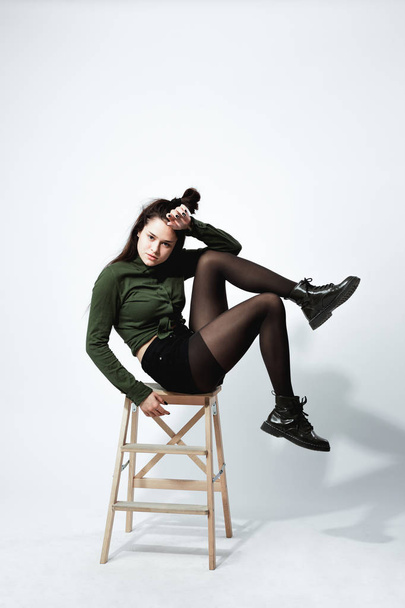 Stylish young dark-haired girl with a funny hairstyle dressed in green shirt, black shorts , tights and shoes is posing sitting on the wooden stool on the white background in the studio - Foto, Bild