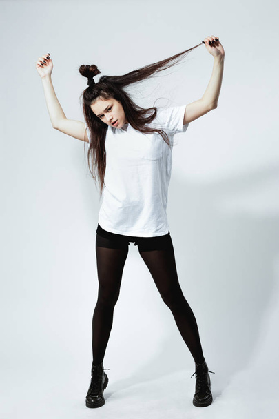 Stylish young dark-haired girl with a funny hairstyle dressed in white t-shirt, black tights and shoes is posing on the white background in the studio - Photo, Image
